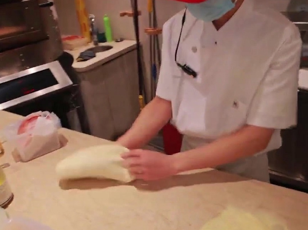 Mani di uno chef che stendono una palla di impasto per pizza su un piano di marmo infarinato.