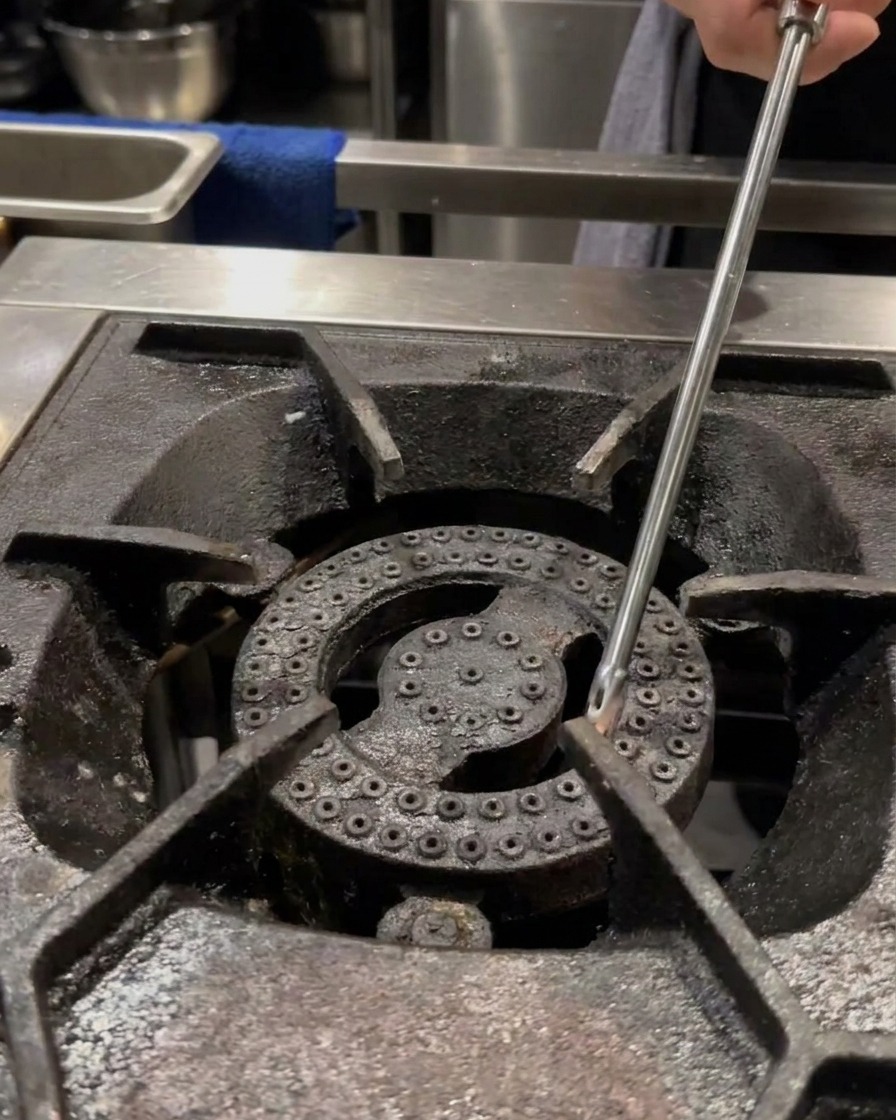 Una mano usa un accendino lungo per accendere un bruciatore a gas professionale con una piccola pentola vuota sulla griglia.