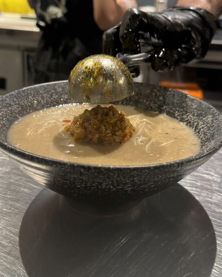 Una mano con guanto che versa olio al peperoncino rosso brillante a cerchi sulla superficie di una ciotola di ramen.