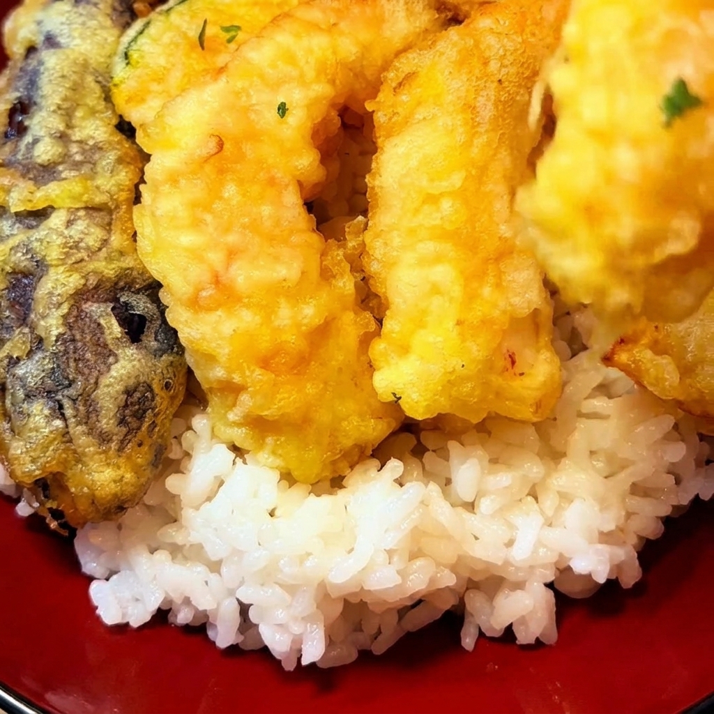 Pezzi di tempura fritta dorata, inclusi gamberi e verdure, disposti sopra un letto di riso bianco in una ciotola rossa.