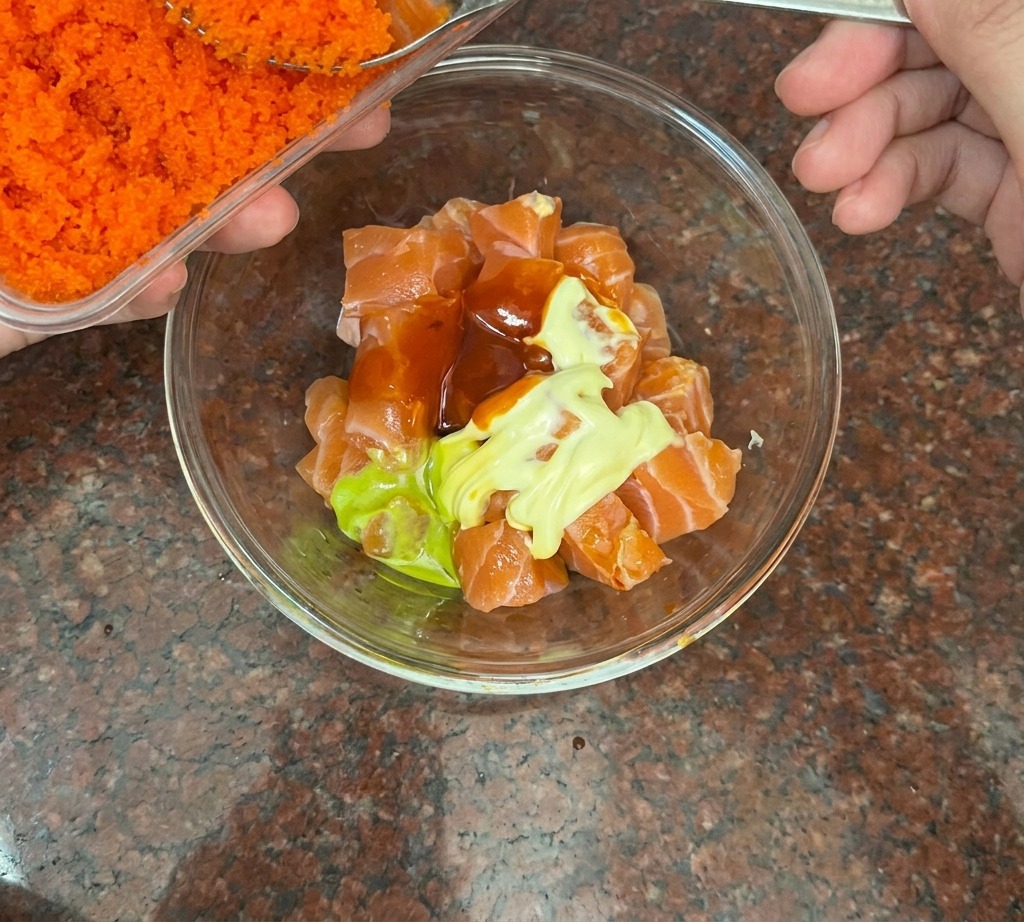 Un cucchiaio dargento aggiunge uova di pesce arancione brillante a una ciotola con salmone a cubetti.