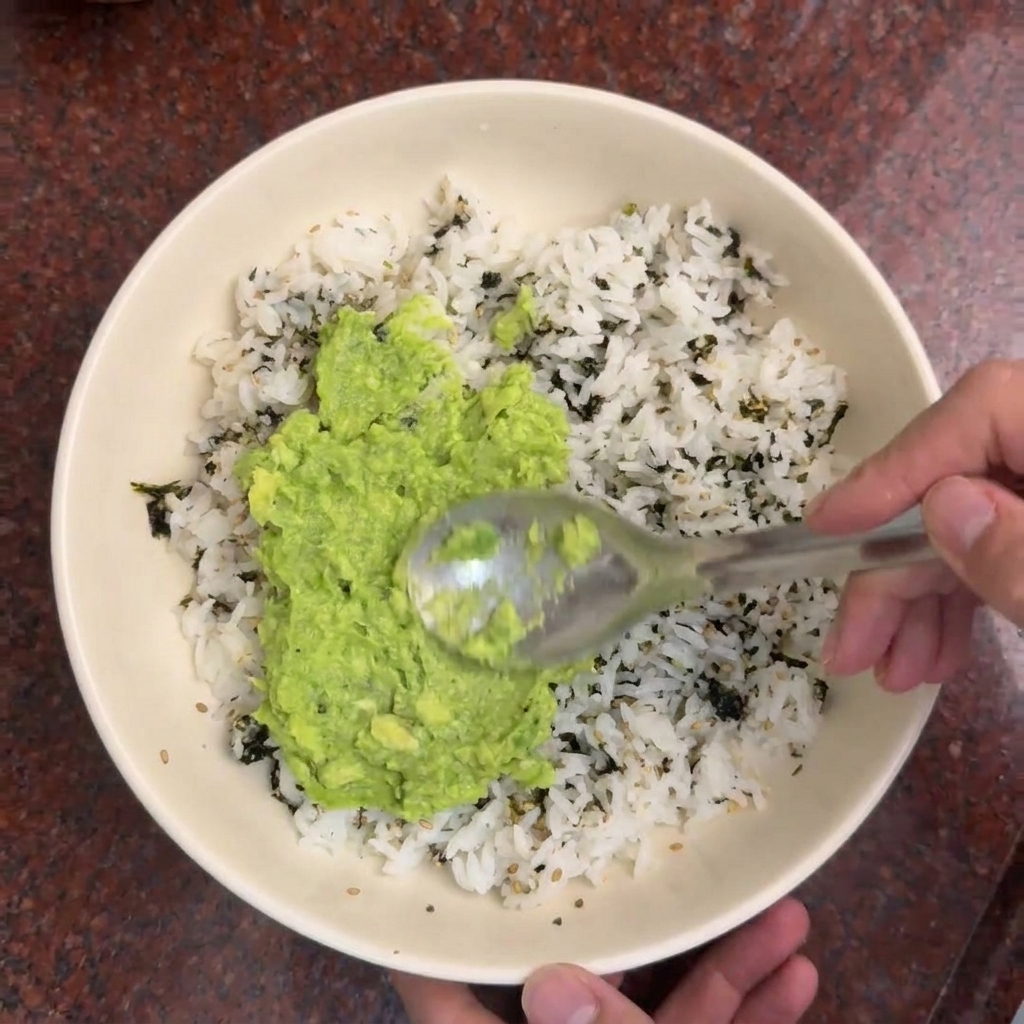 Un cucchiaio stende la purea di avocado verde brillante su una ciotola di riso bianco condito con strisce di alga scura.