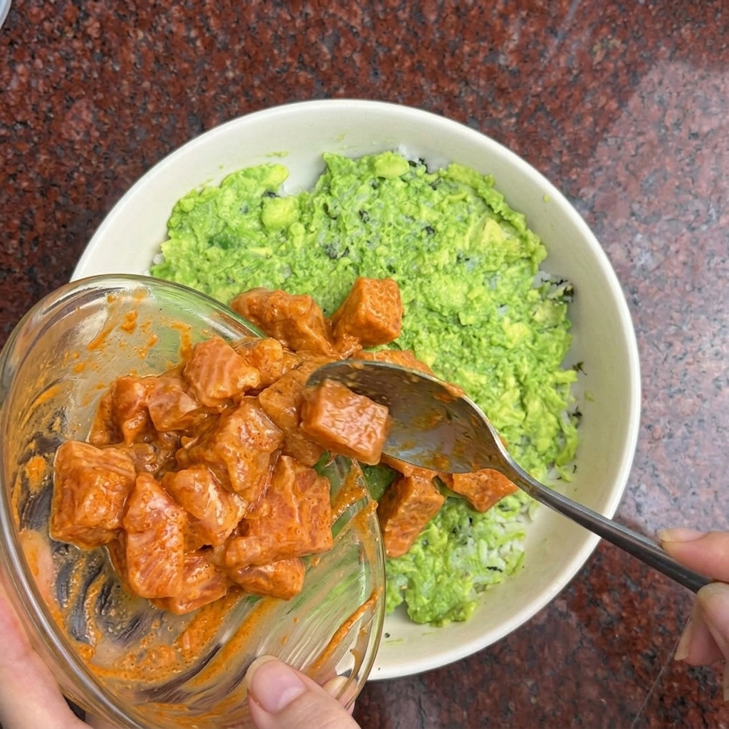 Una mano usa un cucchiaio per disporre i cubetti di salmone marinato sopra la ciotola di riso e avocado.