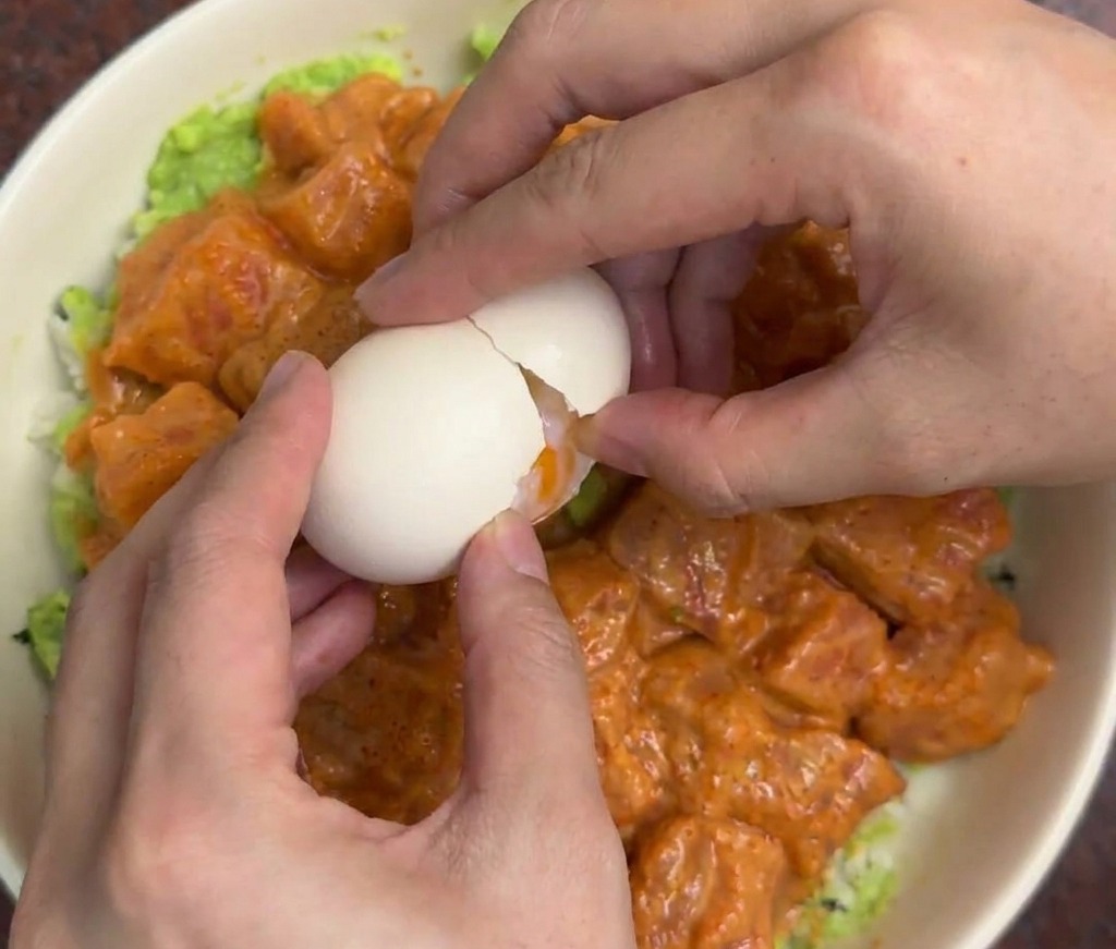 Mani che sgusciano delicatamente un uovo onsen al centro di una ciotola di riso con salmone e avocado.