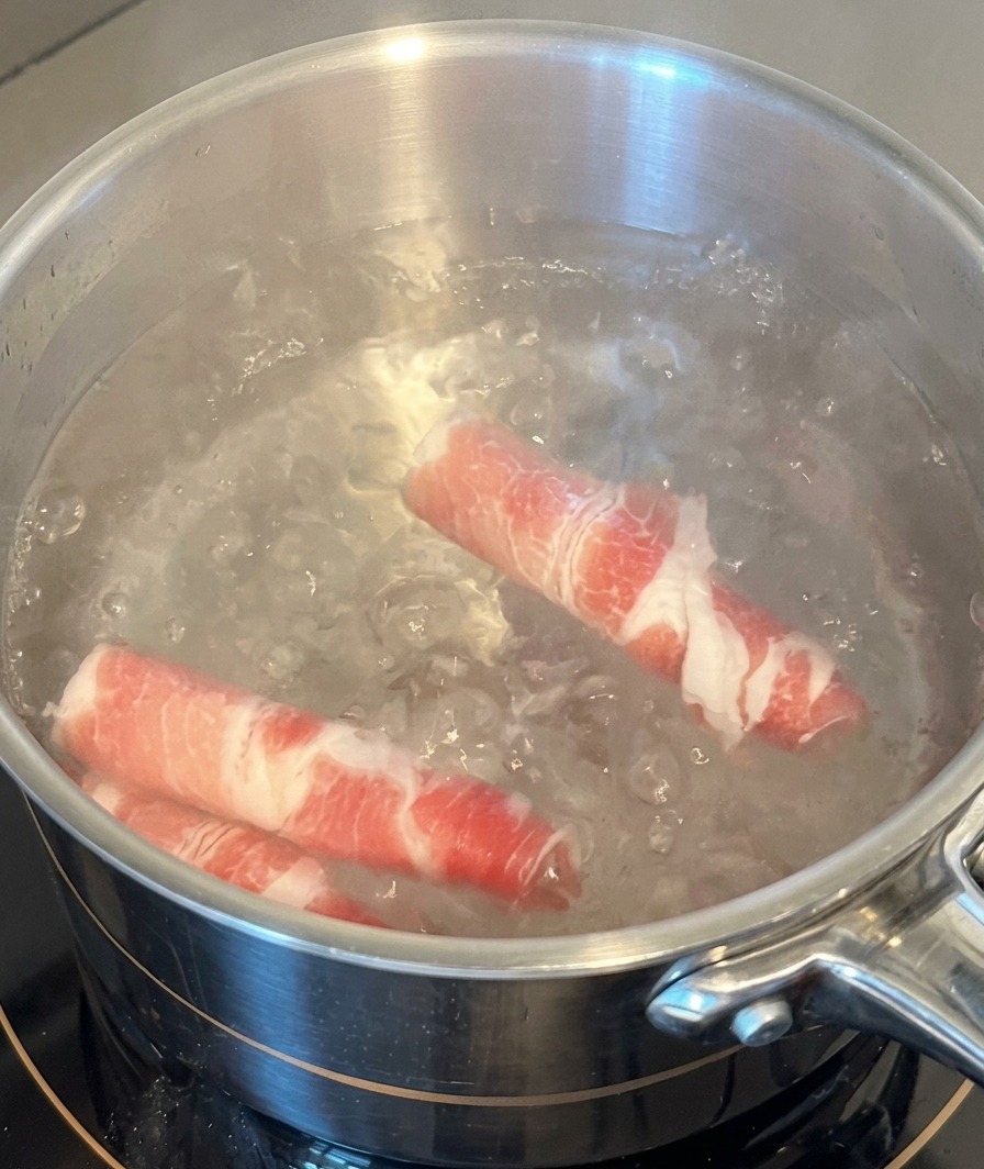 Due rotoli di fette di manzo crudo che vengono immersi in una pentola dacciaio con acqua bollente.