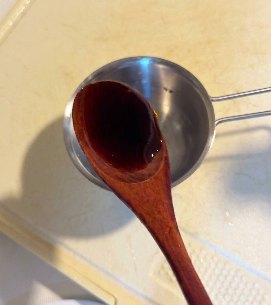Un cucchiaio di legno che versa la scura salsa dostrica in una ciotolina di metallo con gli altri ingredienti.