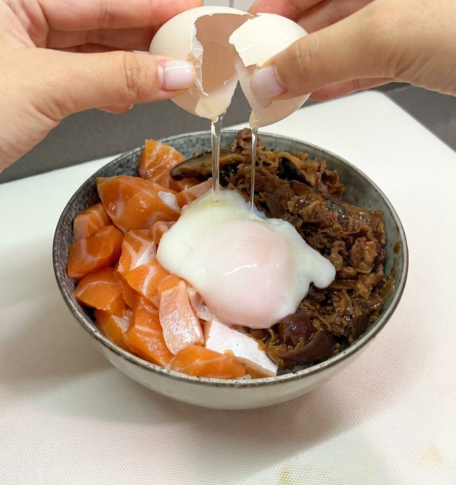 Due mani che aprono delicatamente il guscio di un uovo bianco per far scivolare un uovo onsen morbido su una ciotola di riso con salmone e manzo.