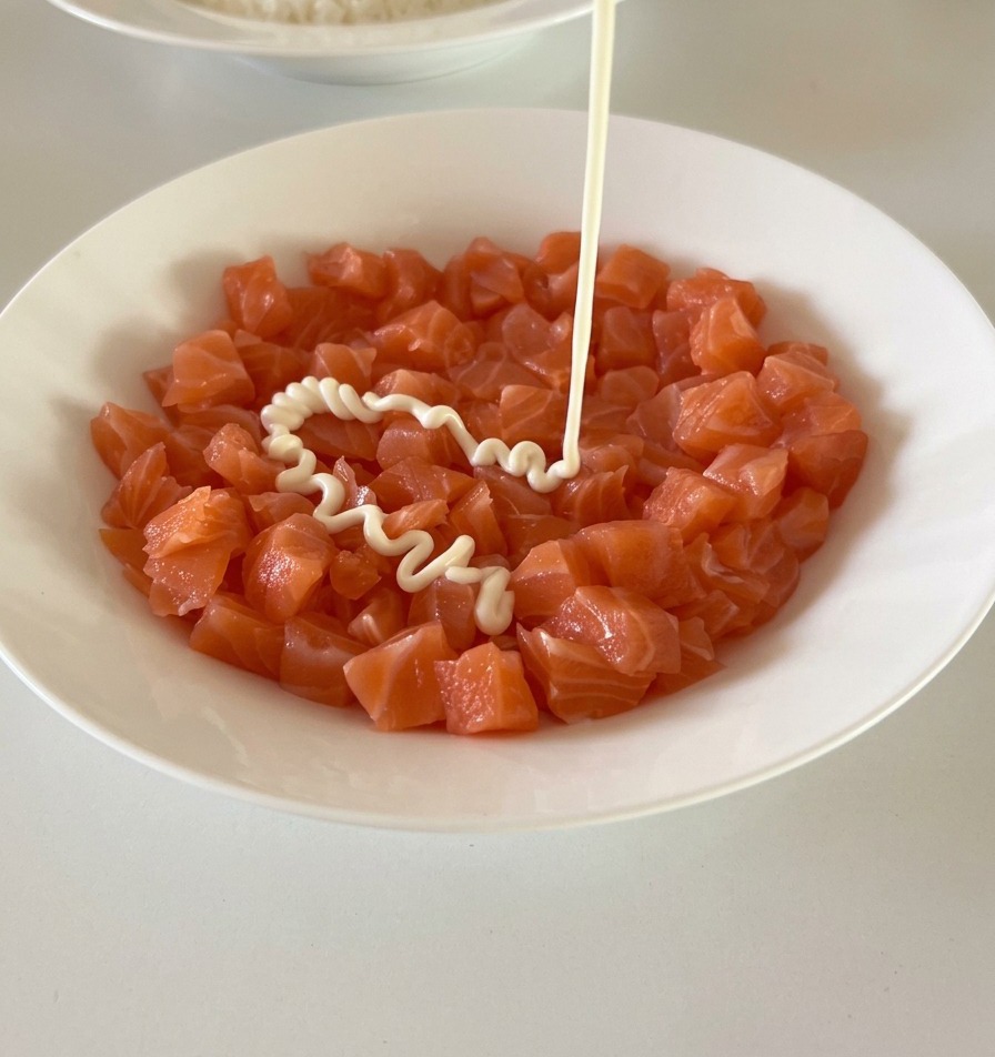Maionese bianca che viene versata da un flacone sopra una ciotola piena di cubetti di salmone.