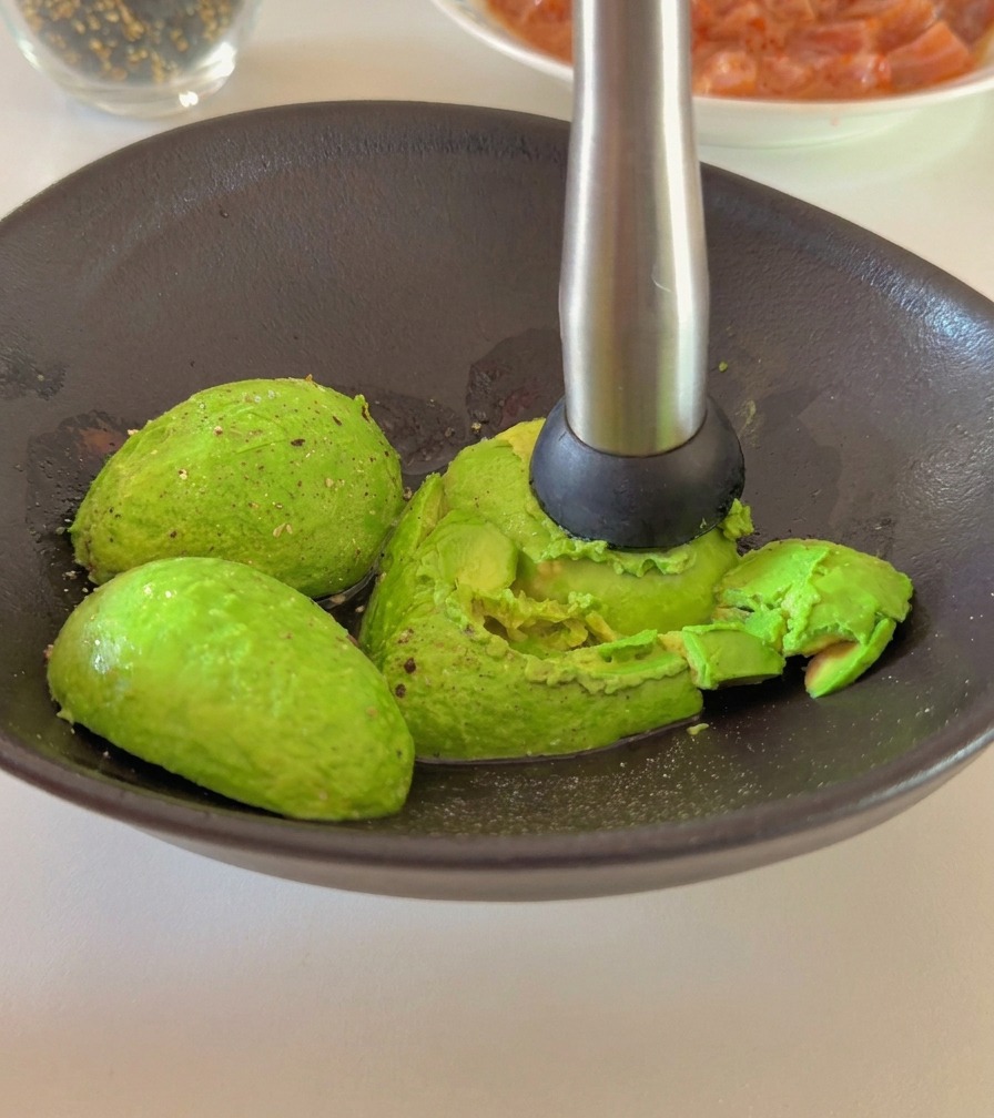 Una persona che usa uno schiacciapatate per ridurre in purea lavocado in una ciotola scura.