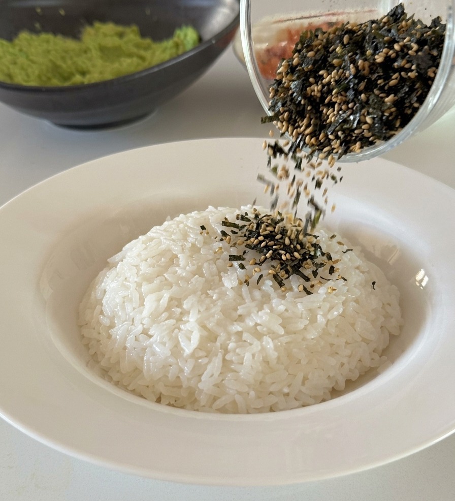 Fiocchi di alga nori e semi di sesamo nero versati da un bicchiere sopra una porzione di riso bianco.