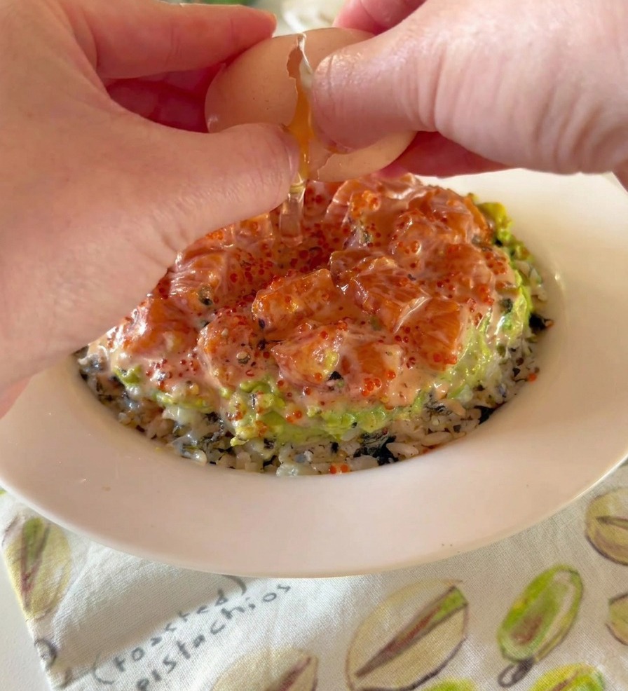 Mani che aprono un uovo onsen morbido sopra una poke bowl stratificata con riso, avocado e salmone.