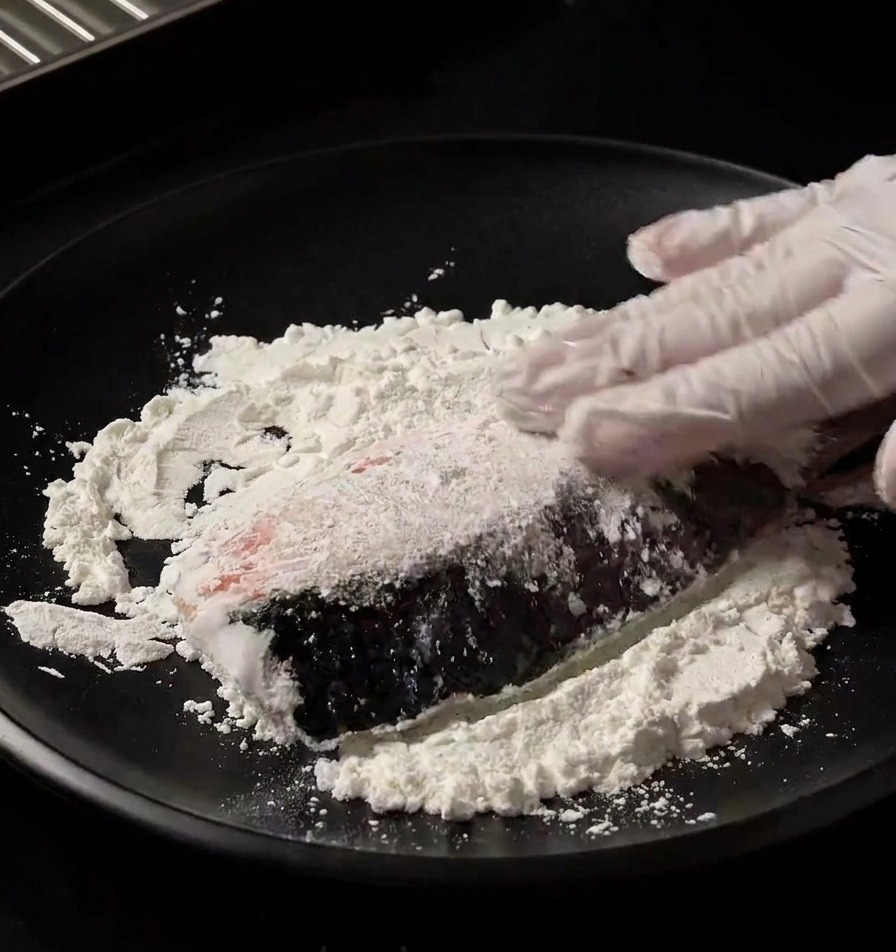 Una mano con un guanto che preme un filetto di salmone fresco su un letto di amido bianco in un piatto nero per impanarlo uniformemente.