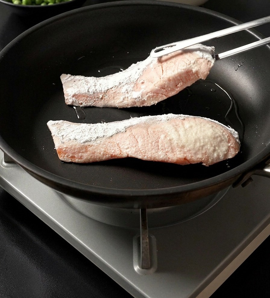 Pinze di metallo che adagiano un filetto di salmone infarinato nellamido in una padella calda accanto a un altro filetto.