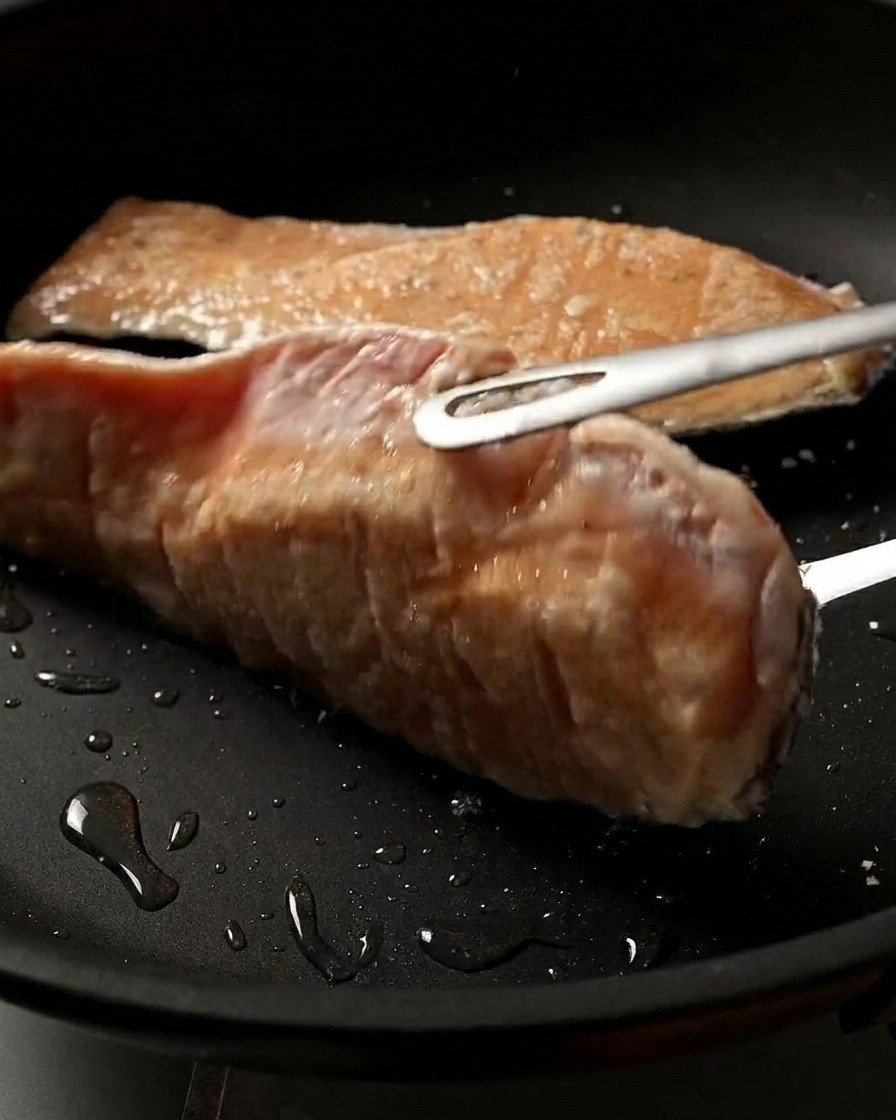 Un primo piano di un filetto di salmone che viene girato in padella, mostrando la sua superficie perfettamente dorata, croccante e ben rosolata.