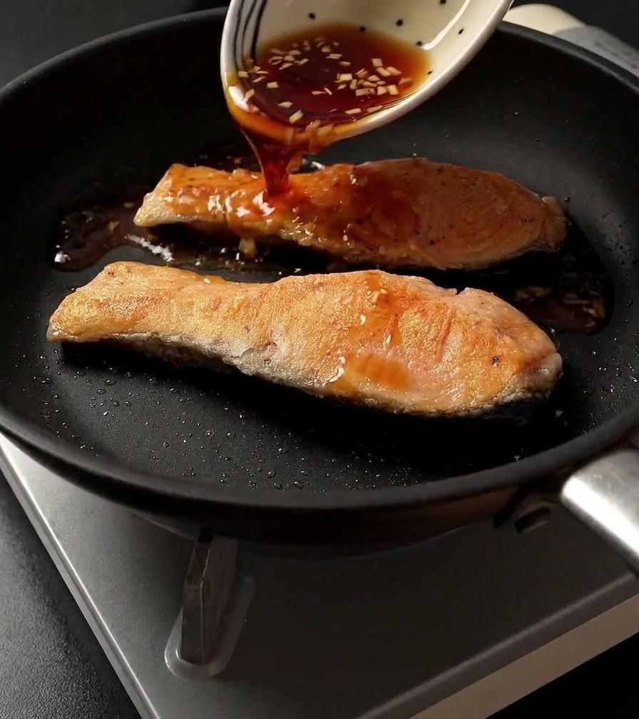 Salsa teriyaki scura versata da una ciotola bianca decorata su due filetti di salmone rosolati in una padella.