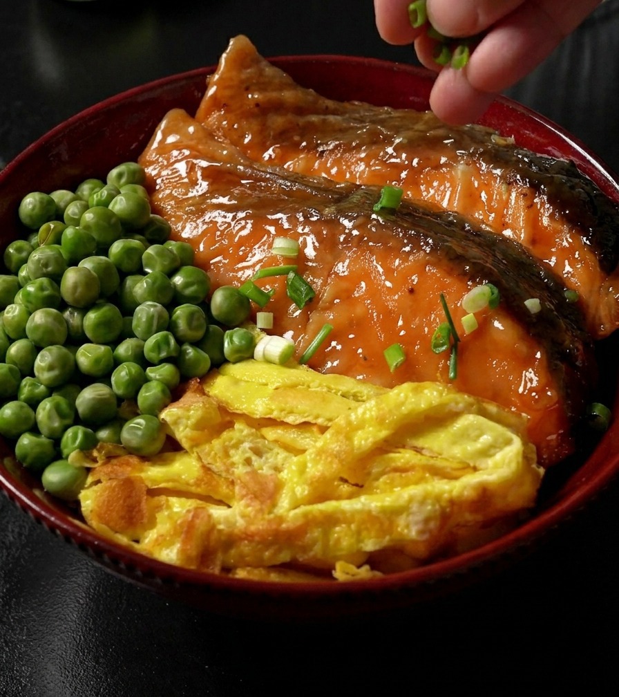 Una mano che spolvera cipollotti verdi affettati su una ciotola di riso completa, guarnita con salmone glassato teriyaki, striscioline duovo e piselli verdi.