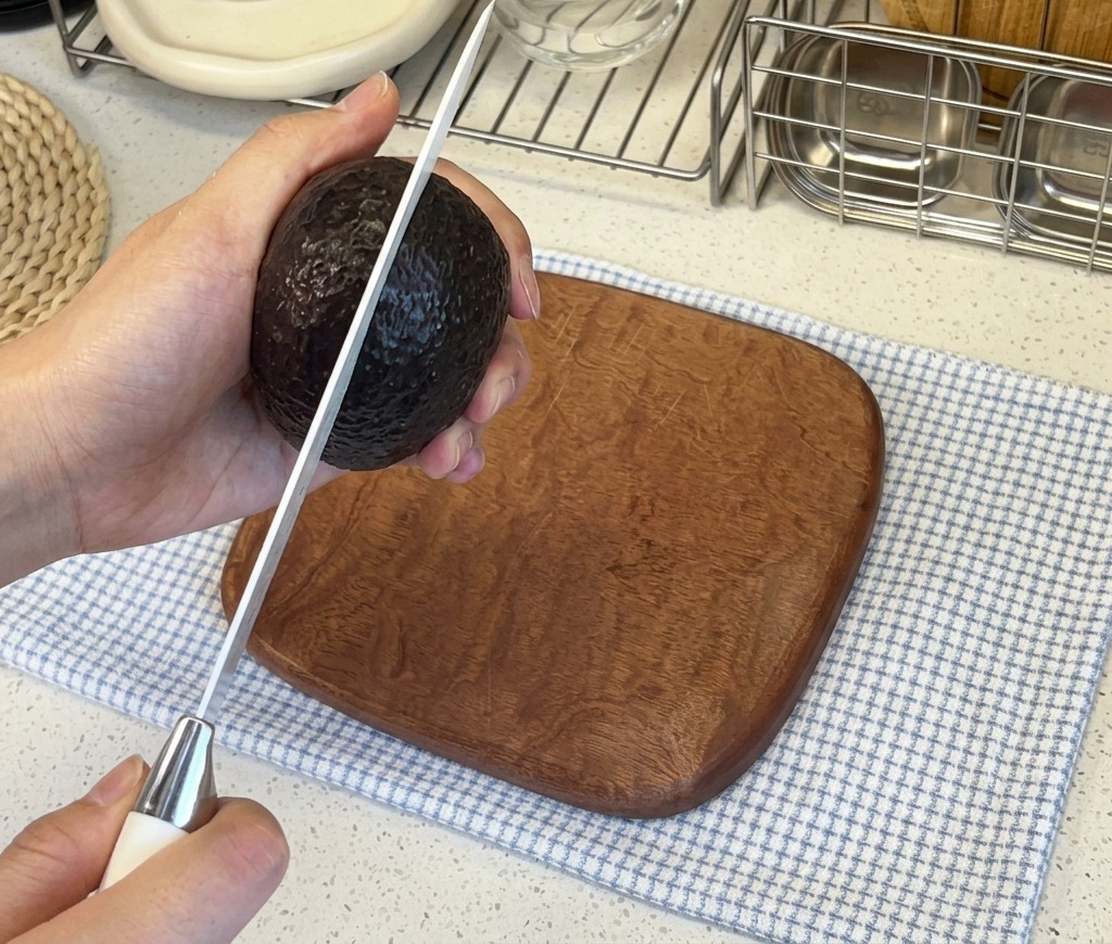 Mani che tengono un avocado dalla buccia scura mentre lo tagliano verticalmente con un coltello da cucina affilato.