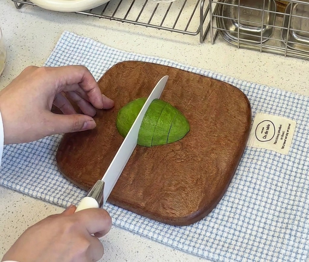 Una mano che tiene ferma metà di un avocado sbucciato su un tagliere mentre un coltello bianco lo taglia a piccoli cubetti.
