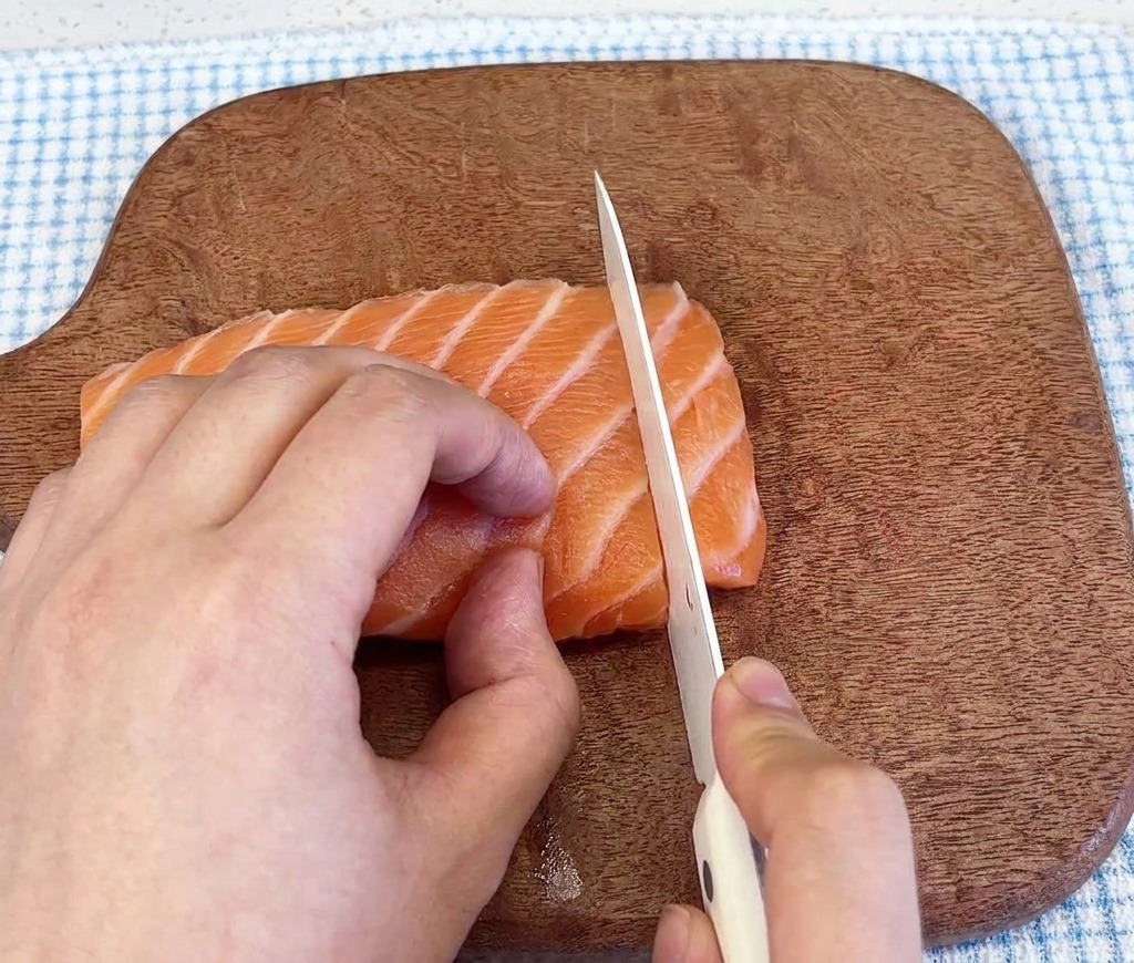 Primo piano di un coltello affilato che taglia un filetto di salmone fresco e venato su un tagliere di legno scuro.
