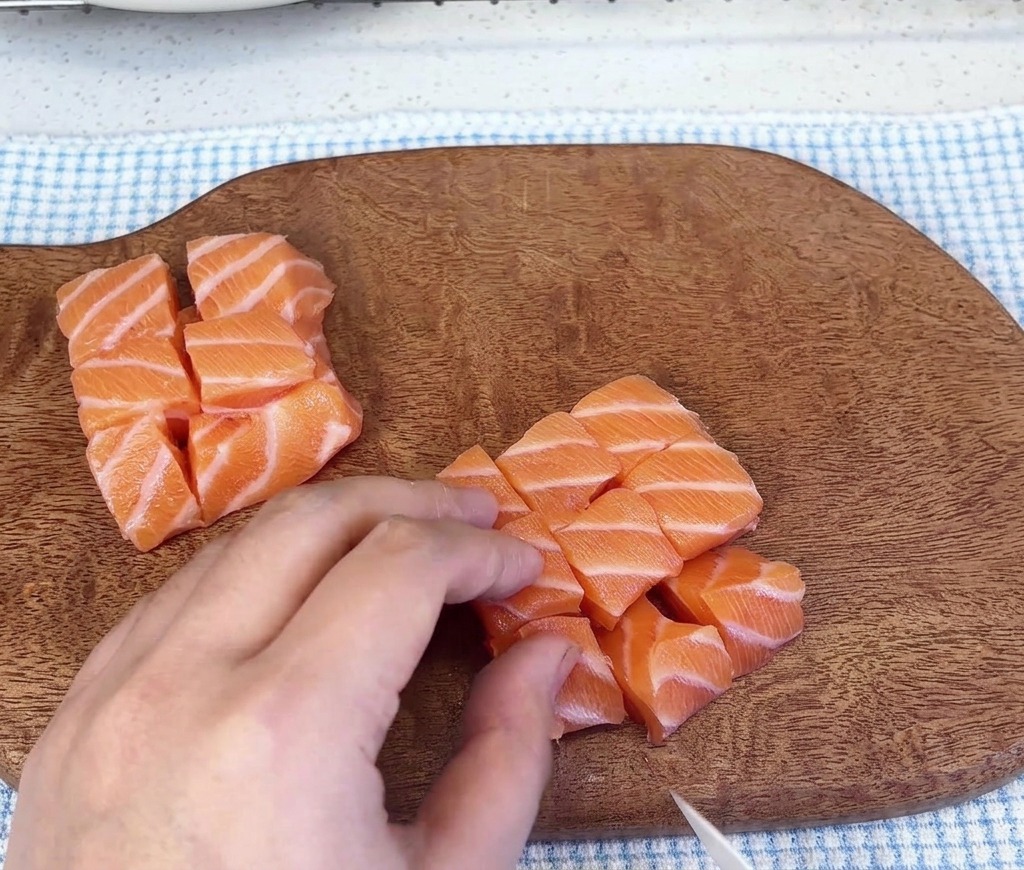 Piccoli mucchietti di salmone crudo tagliato a cubetti adagiati su un tagliere di legno dopo il taglio.
