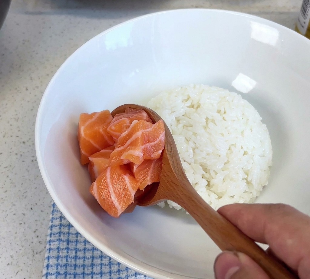 Un cucchiaio di legno che solleva una porzione di cubetti di salmone fresco per adagiarli in una ciotola bianca con riso al vapore.