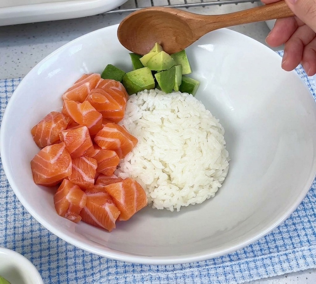 Cubetti di avocado che vengono versati con un cucchiaio in una ciotola bianca accanto al salmone crudo sopra il riso.