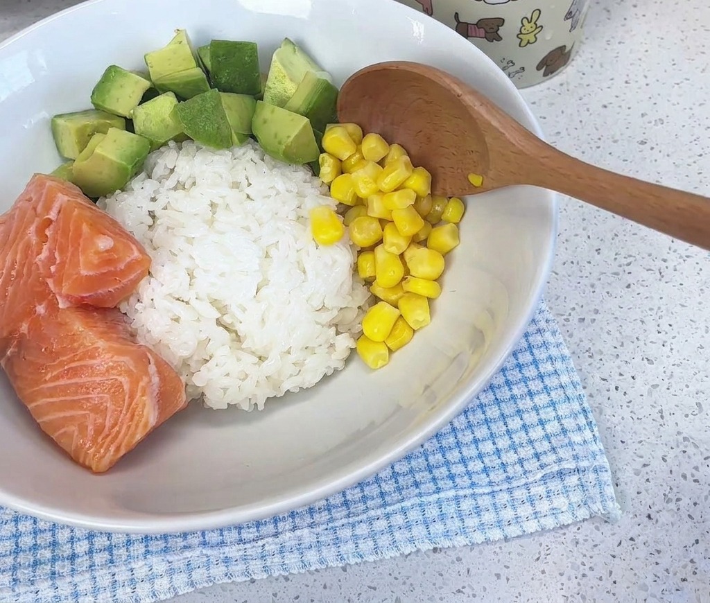 Un cucchiaio di legno che aggiunge chicchi di mais giallo brillante in una ciotola bianca contenente riso, avocado a cubetti e pezzi di salmone fresco.