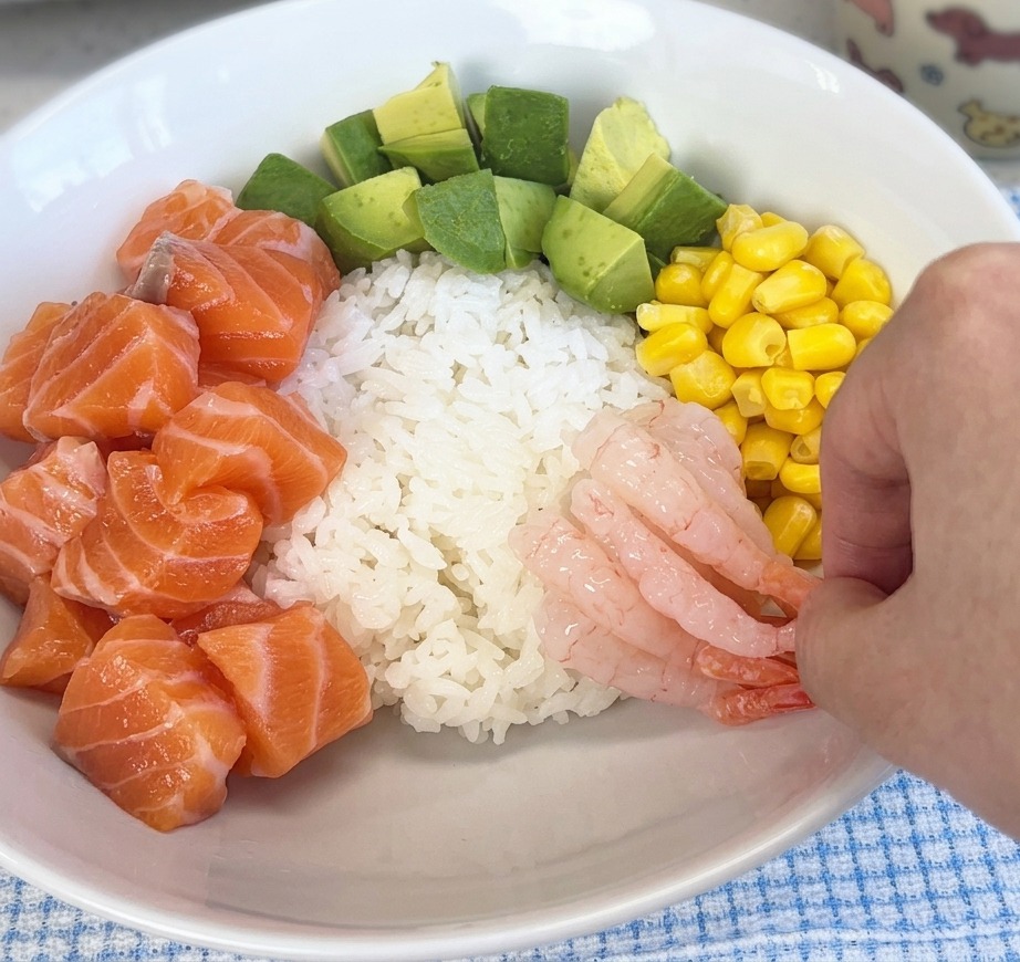 Una mano che adagia gamberetti dolci, rosa e traslucidi, sul riso allinterno di una ciotola insieme a salmone e mais.