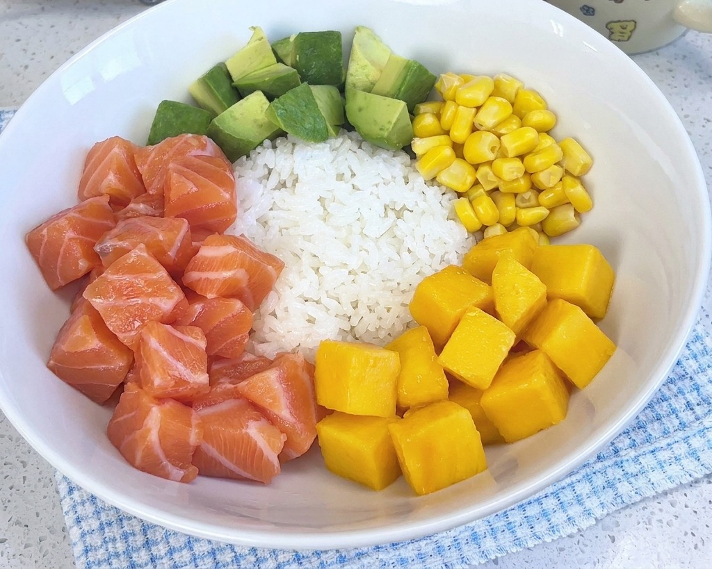 Una poke bowl colorata con riso bianco, salmone, avocado, mais e gamberetti, che ora include anche un mucchietto di cubetti di mango giallo brillante.