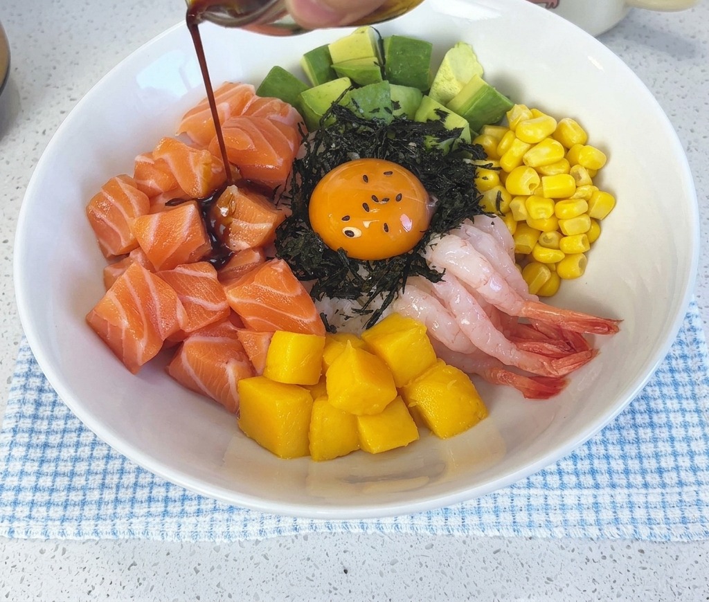 Salsa di soia scura al wasabi che viene versata da una piccola ciotola di vetro sulla poke bowl al salmone già assemblata e sul tuorlo duovo.
