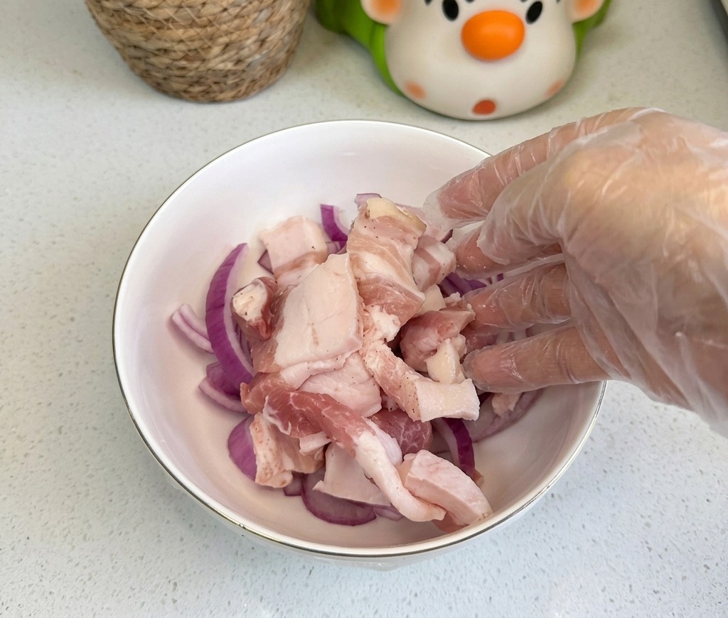 Fette di pancetta di maiale cruda aggiunte a una ciotola bianca contenente cipolle rosse affettate sottilmente.