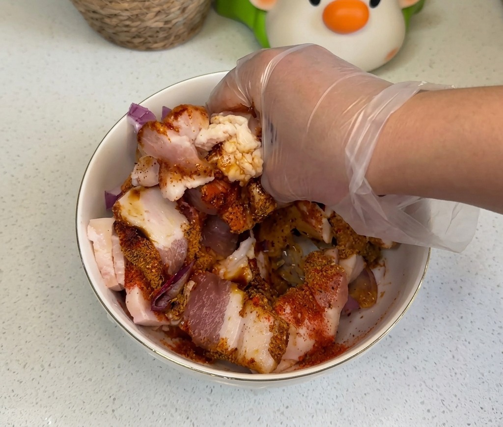 Una mano con un guanto di plastica trasparente che mescola accuratamente le fette di pancetta cruda con cipolle e spezie colorate in una ciotola.