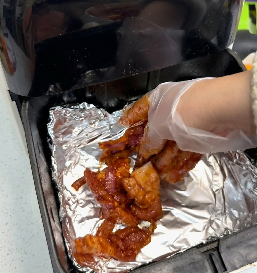 Una mano con un guanto trasparente che dispone le fette di pancetta marinate su un foglio di alluminio argentato dentro il cestello nero della friggitrice ad aria.