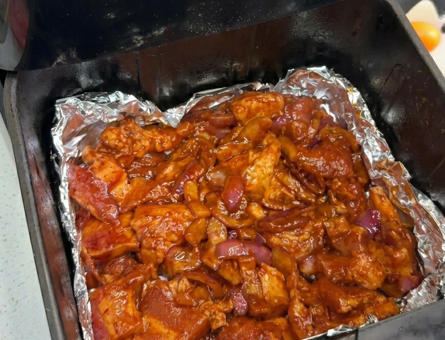 Pancetta marinata e cipolle rosse distribuite uniformemente sul fondo di un cestello della friggitrice ad aria rivestito di alluminio.