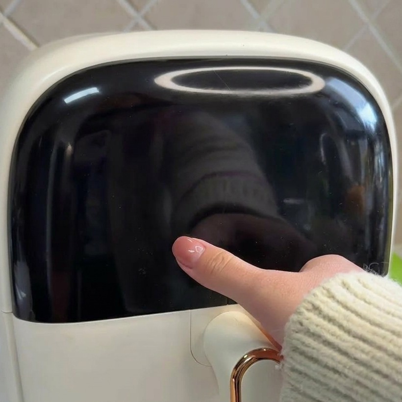 Primo piano di una mano che tocca il display digitale nero di una friggitrice ad aria bianca.
