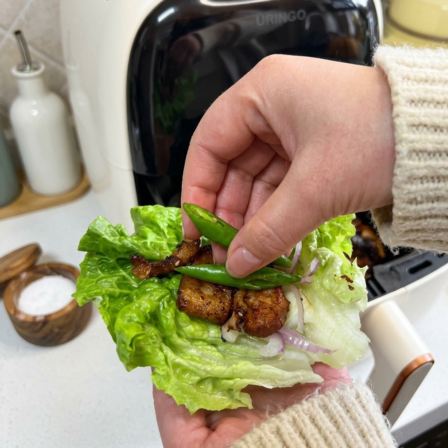 Una mano che posiziona due piccoli peperoncini verdi brillanti su un involtino di lattuga contenente pancetta croccante e cipolle.