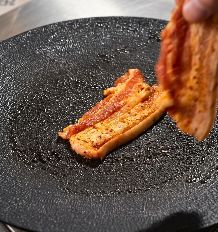Una mano adagia una fetta di pancetta di maiale su una piastra circolare nera.