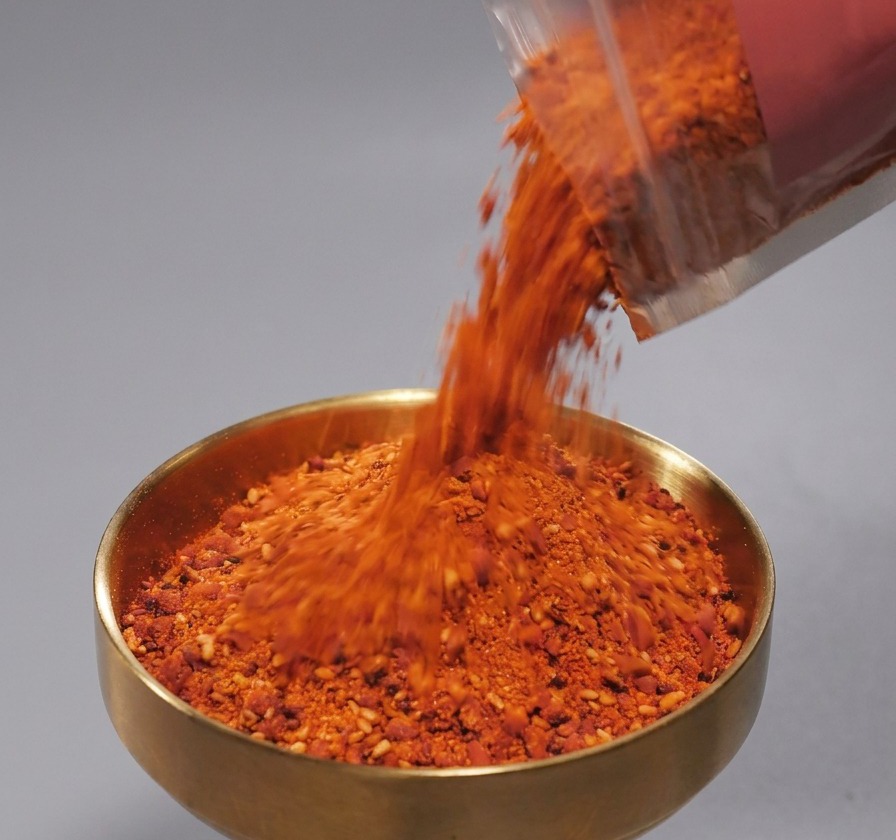 Spezie rosse in polvere con semi di sesamo versate da una bustina in una ciotolina dorata.