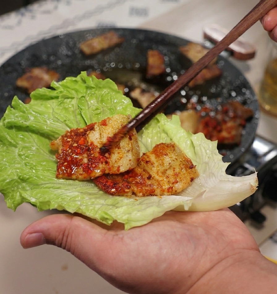 Una mano che tiene una grande foglia di lattuga verde mentre vi adagia sopra un pezzo di pancetta con le bacchette.
