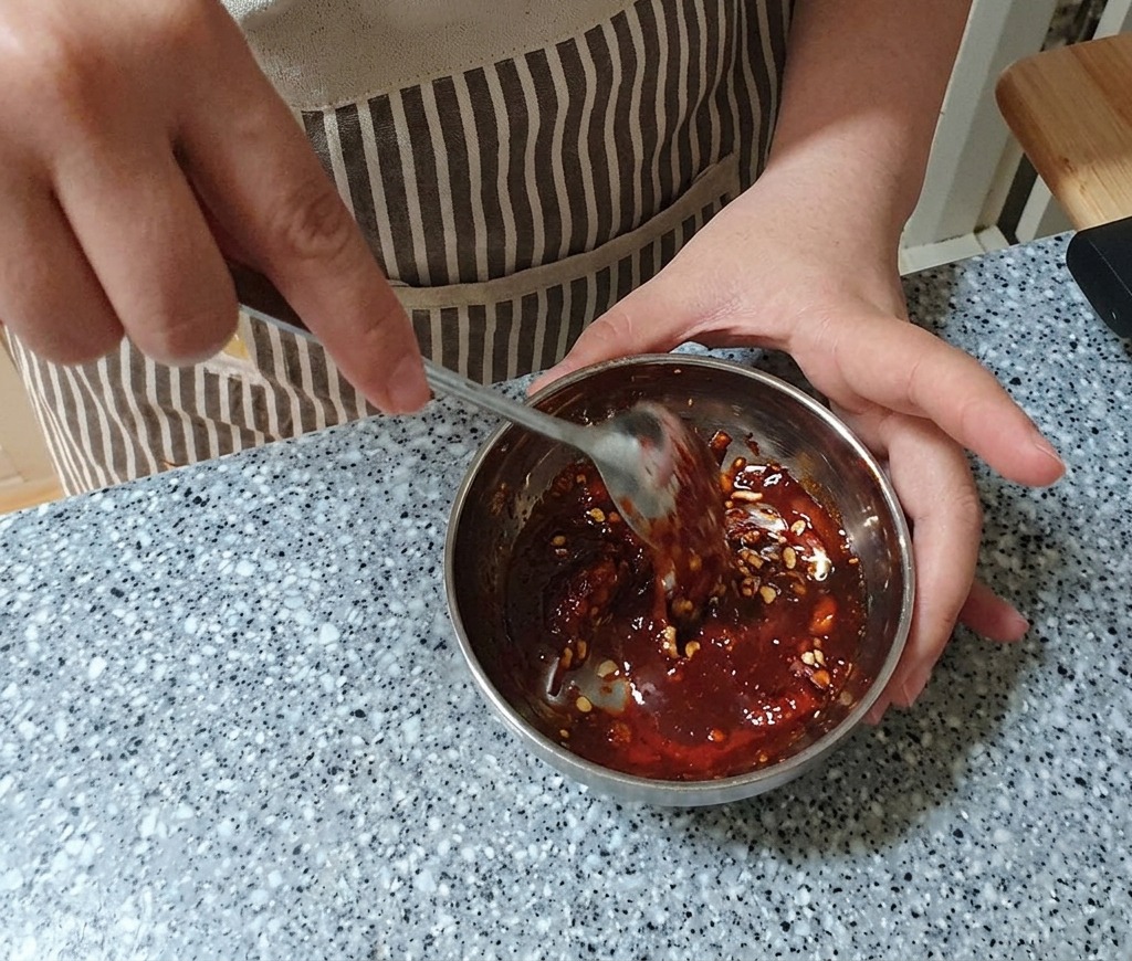 Una marinatura densa e di colore rosso scuro che viene mescolata in una ciotola di metallo con un cucchiaio in acciaio inossidabile fino a risultare completamente omogenea.