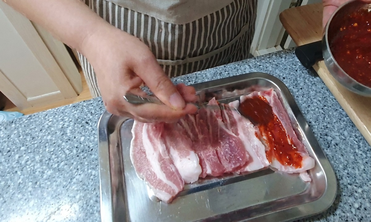 Una mano che usa un cucchiaio per rivestire le strisce di pancetta di maiale cruda con una densa e vivace marinatura al peperoncino rosso su un vassoio dargento.