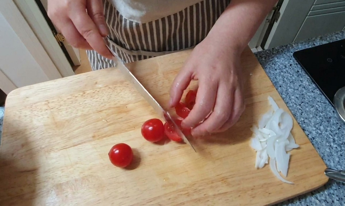 Piccoli pomodorini rossi tagliati a metà su un tagliere di legno accanto a un mucchietto di cipolle bianche affettate.