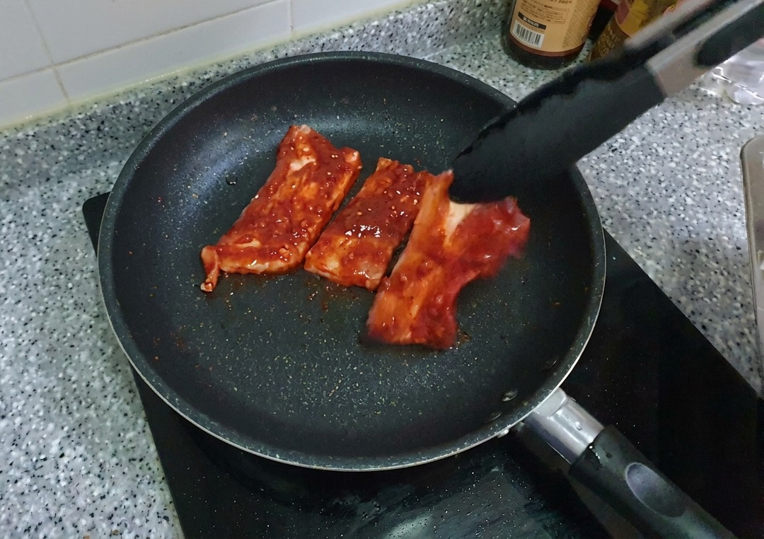 Strisce di pancetta di maiale rossa e marinata che vengono adagiate in una padella antiaderente nera usando delle pinze.