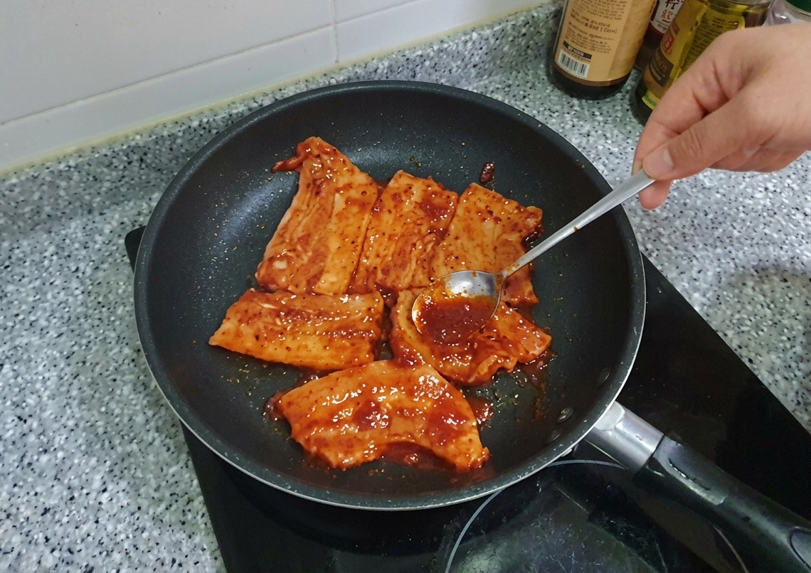 Un cucchiaio che spalma una densa marinata al Gochujang rosso sulle fette di pancetta di maiale in padella.