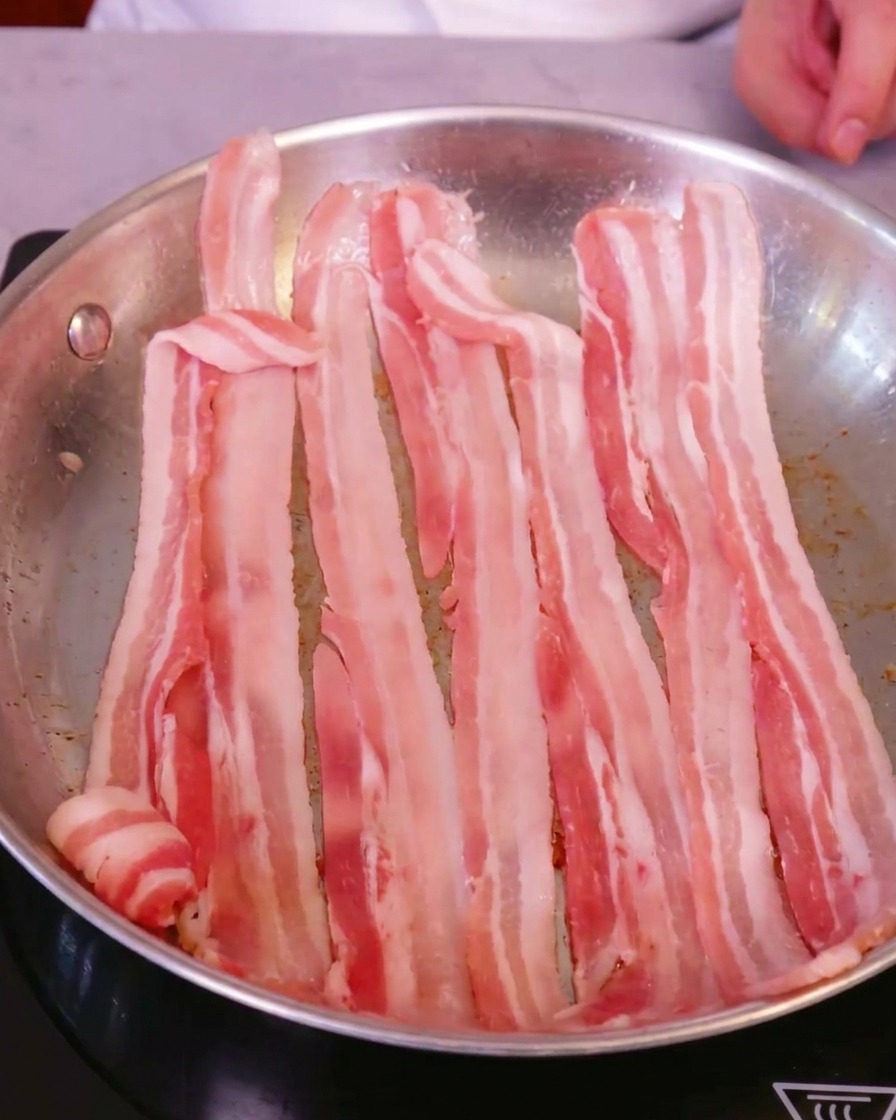 Fette di pancetta di maiale fresca disposte con cura in una grande padella in acciaio inossidabile su un piano a induzione.