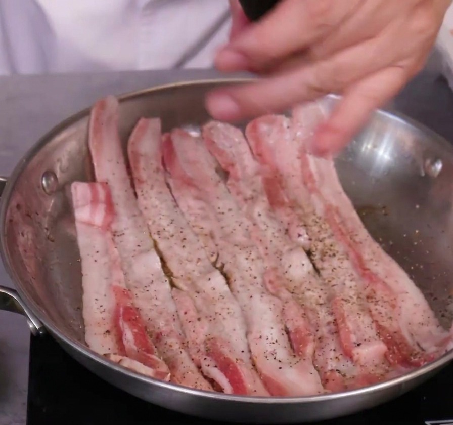 Fette di pancetta di maiale cruda condite con un macinapepe nero manuale in una padella di acciaio inossidabile.