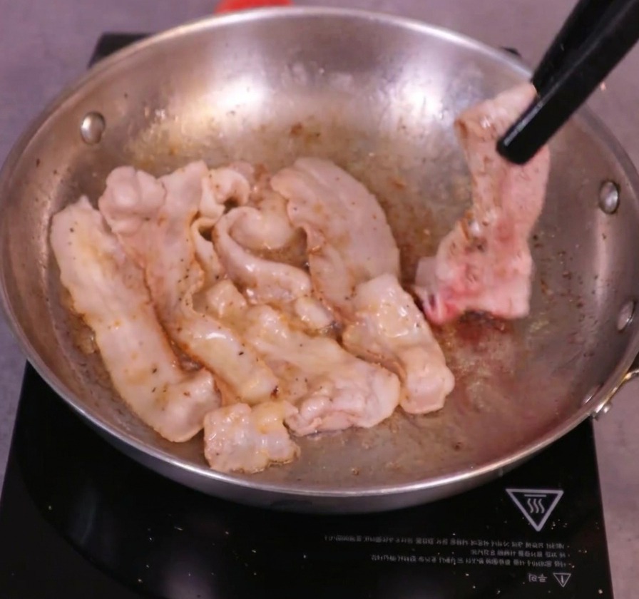 Fette di pancetta dorate e sfrigolanti girate in padella usando delle pinze da cucina nere.