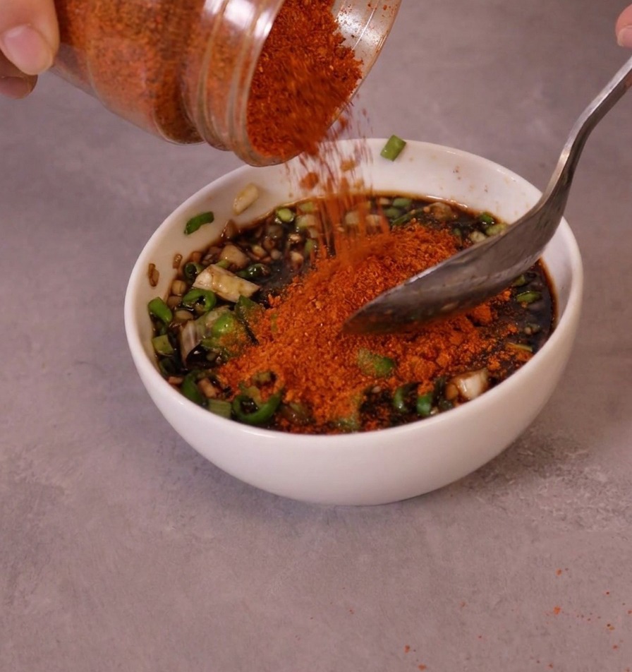 Peperoncino in polvere rosso brillante versato da un barattolo in una ciotola con la base della salsa e i peperoncini verdi.