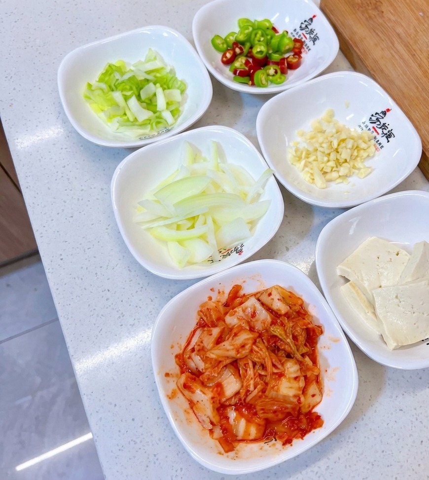 Una selezione di ciotoline bianche contenenti cipollotti tritati, peperoncini verdi e rossi, aglio tritato, cipolla affettata, tofu e kimchi.
