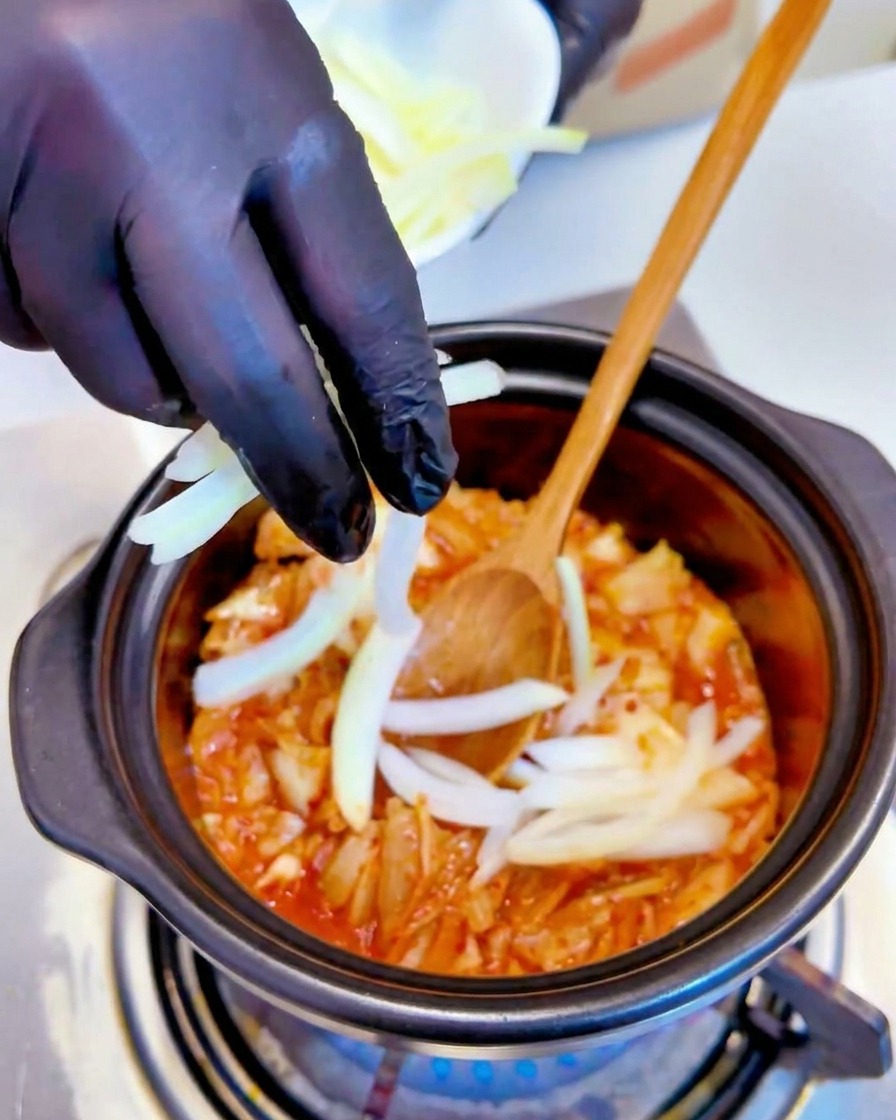 Cipolle bianche affettate vengono aggiunte al composto di kimchi che sobbolle in una pentola da una mano con un guanto nero.